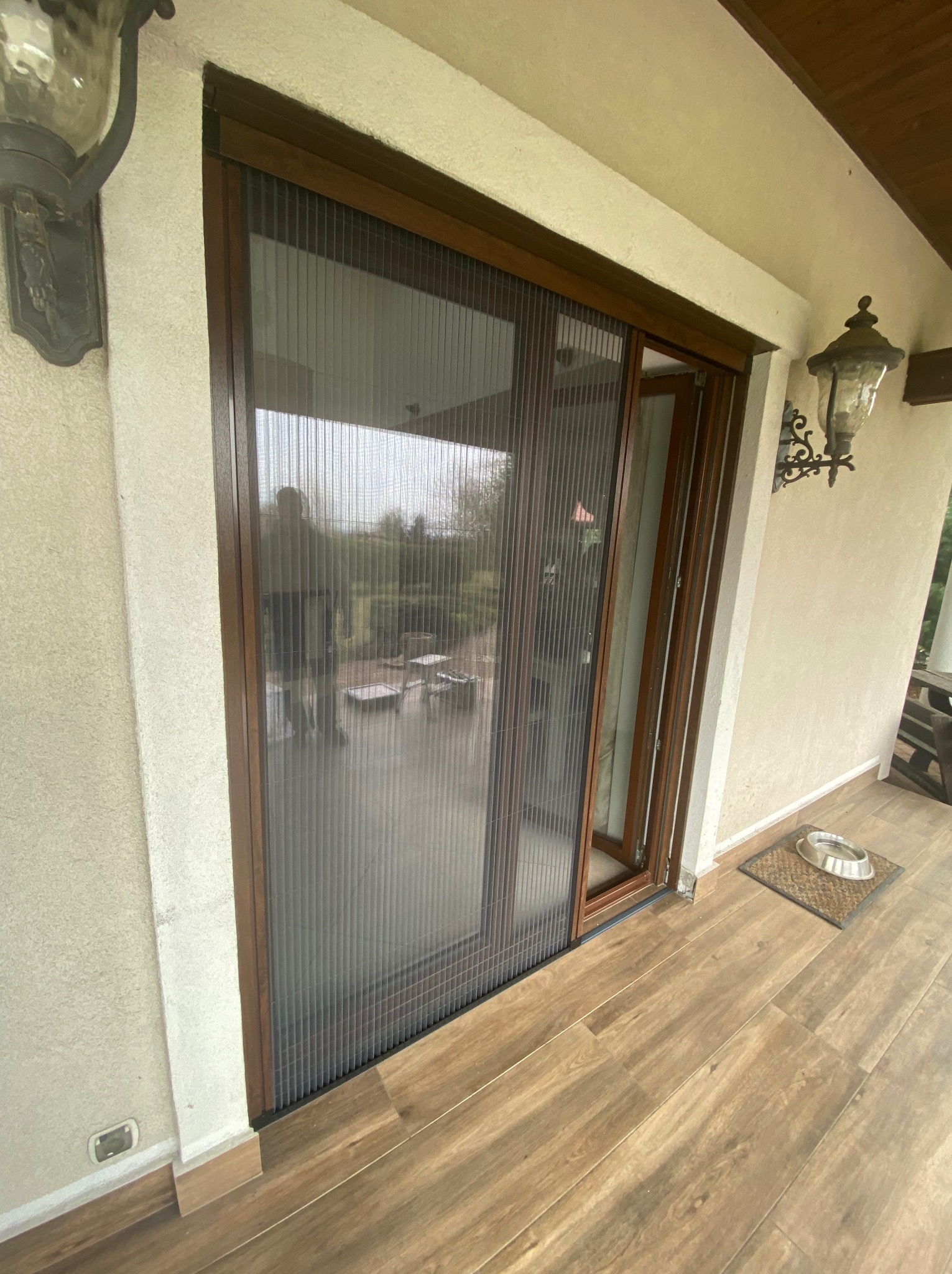 Zewnętrzna moskitiera plisowana w brązowej ramie, zamontowana na tarasie z drewnopodobną podłogą i widoczną lampą ścienną.