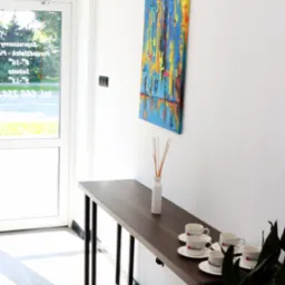 Jasne wnętrze biura z minimalistycznym wystrojem: ciemny stół z czterema filiżankami z logo firmy ubezpieczeniowej, obraz z motywem deszczu i parasoli, białe ściany, klimatyzator oraz roślina...