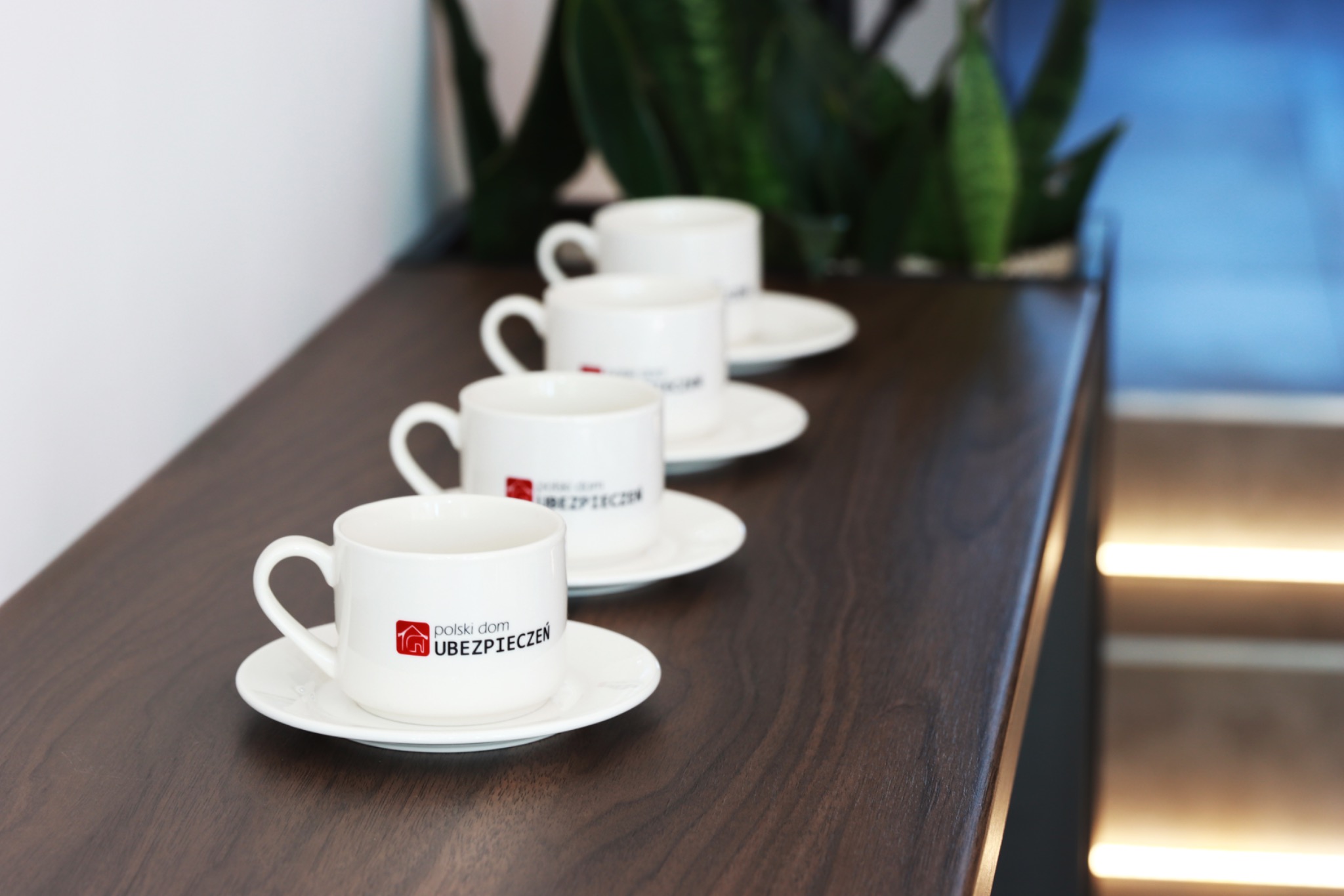 Cztery białe filiżanki z logo firmy ubezpieczeniowej ustawione w rzędzie na ciemnym, drewnianym blacie, z rośliną w tle.