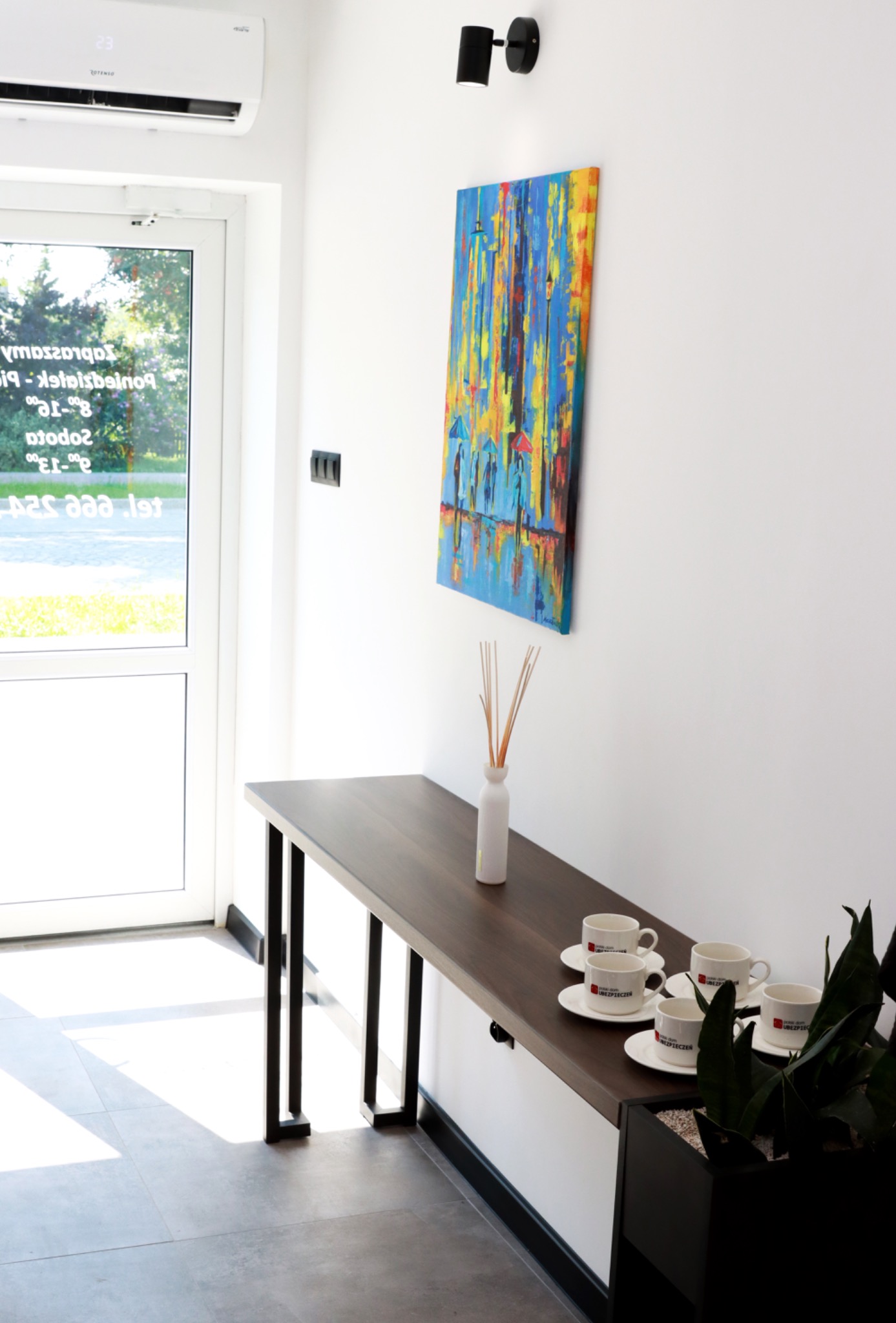 Jasne wnętrze biura z minimalistycznym wystrojem: ciemny stół z czterema filiżankami z logo firmy ubezpieczeniowej, obraz z motywem deszczu i parasoli, białe ściany, klimatyzator oraz roślina...