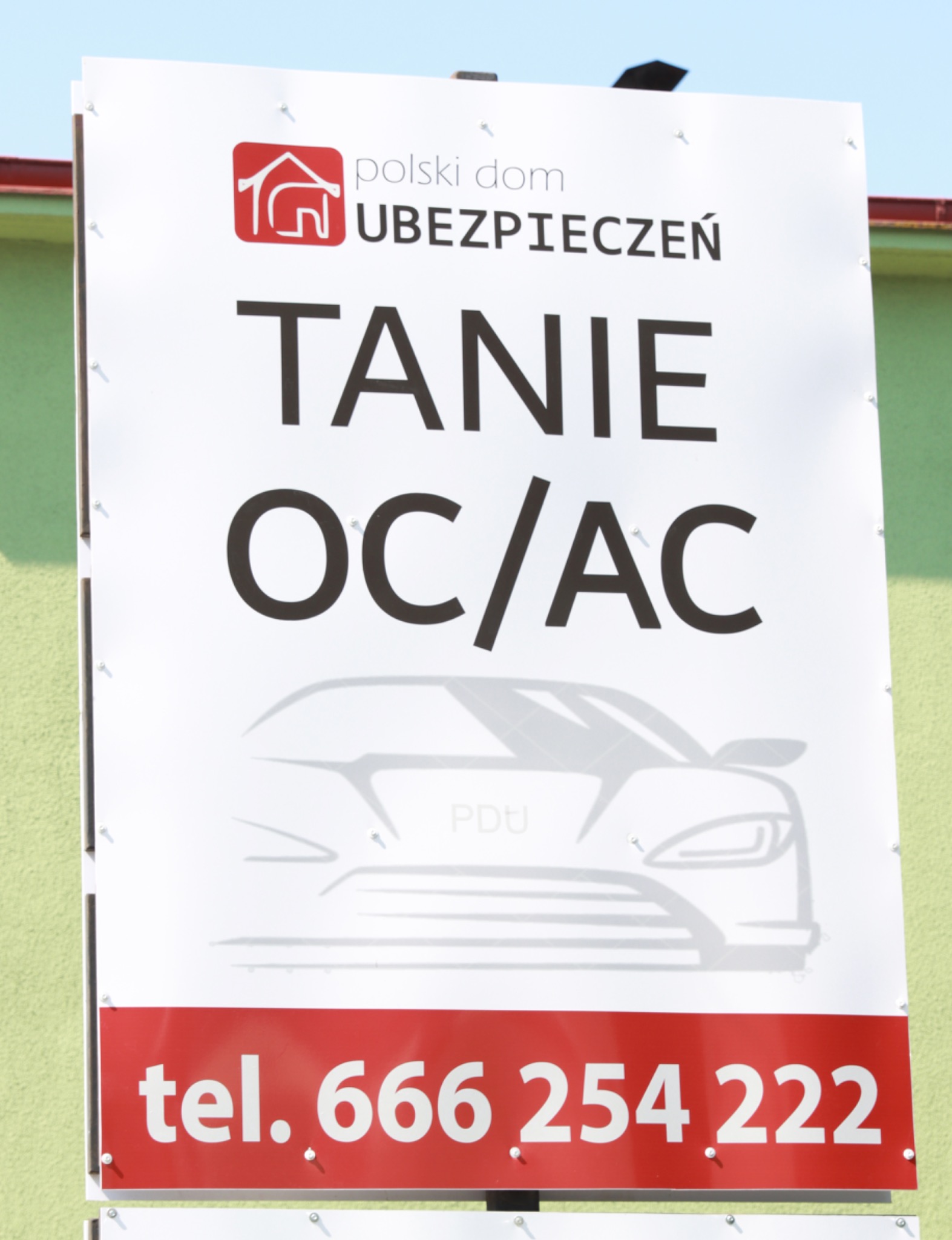 Billboard reklamujący tanie ubezpieczenia OC/AC z logo 'polski dom UBEZPIECZEŃ' i numerem telefonu na tle zielonej ściany.