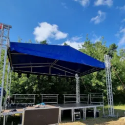 Scena plenerowa z zadaszeniem w kolorze niebieskim, konstrukcja z metalowych kratownic, widoczne elementy systemu nagłośnieniowego i oświetleniowego, tło stanowią drzewa i błękitne niebo z chmurami.