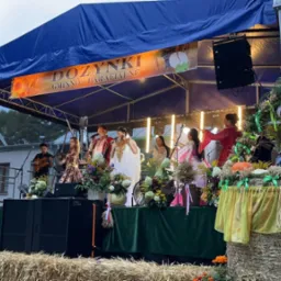 Zespół muzyczny w ludowych strojach występuje na scenie podczas dożynek gminno-parafialnych, scena udekorowana kwiatami i dyniami, nagłośnienie widoczne po bokach.