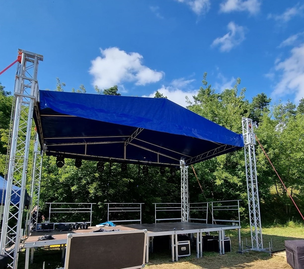 Scena plenerowa z zadaszeniem w kolorze niebieskim, konstrukcja z metalowych kratownic, widoczne elementy systemu nagłośnieniowego i oświetleniowego, tło stanowią drzewa i błękitne niebo z chmurami.