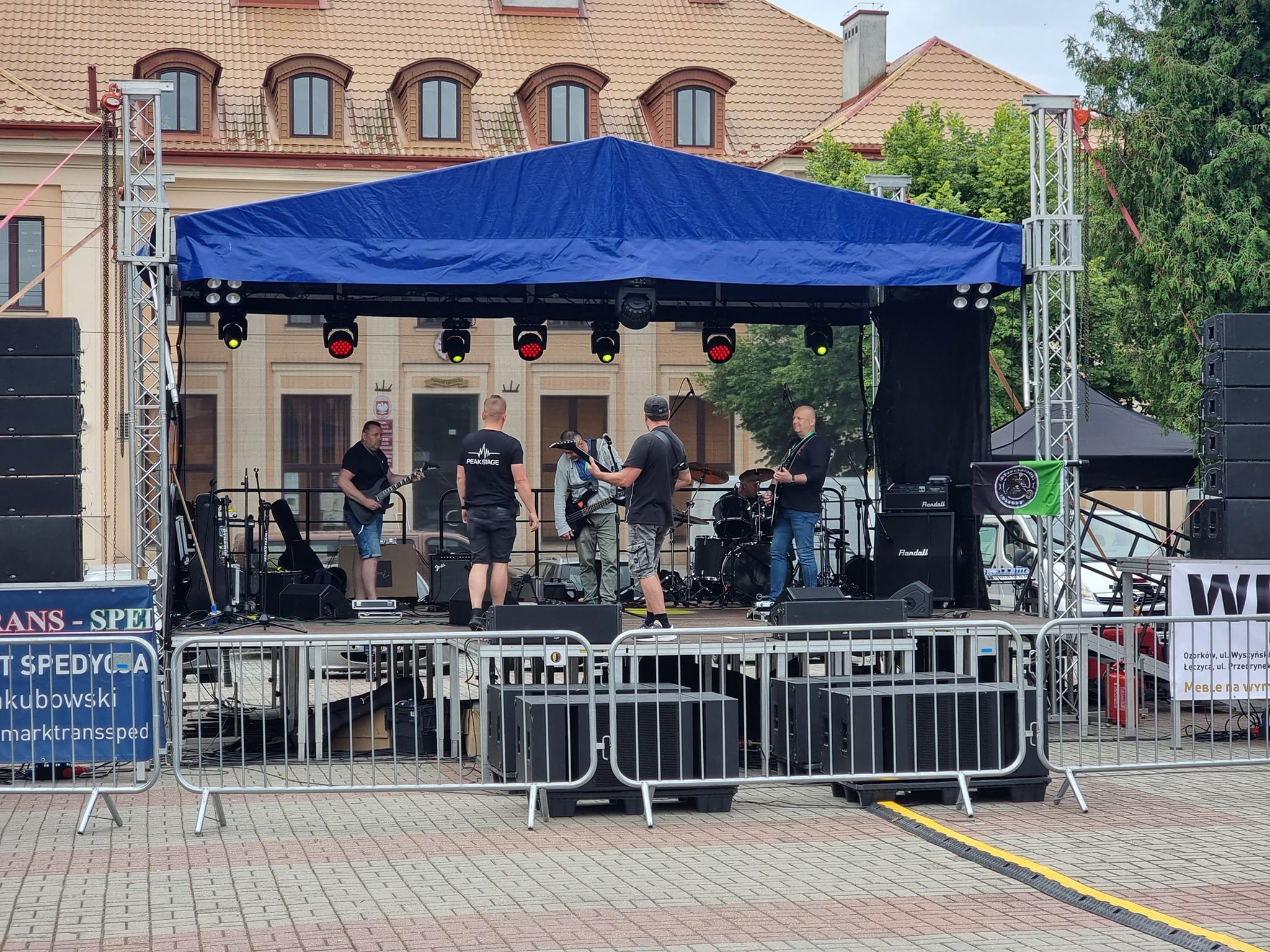 Zespół muzyczny gra koncert na zewnątrz, na scenie z niebieskim zadaszeniem i oświetleniem. Widoczne kolumny głośnikowe po bokach sceny i barierki zabezpieczające. W tle budynek z oknami na poddaszu.