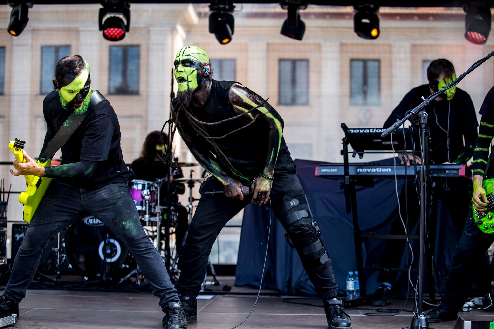 Zespół muzyczny w makijażu scenicznym, grający koncert na scenie z oświetleniem punktowym i budynkiem w tle.