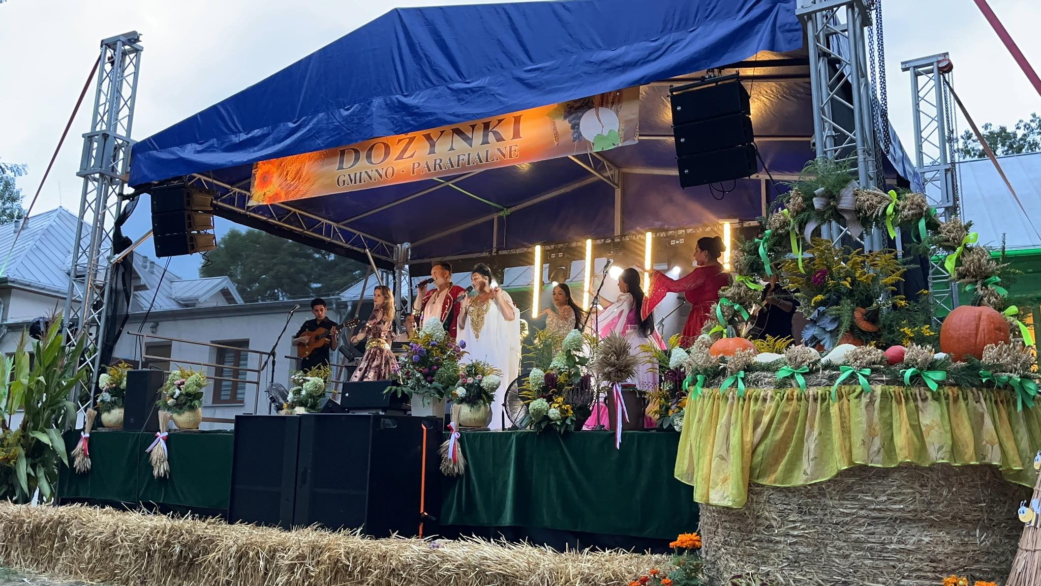 Zespół muzyczny w ludowych strojach występuje na scenie podczas dożynek gminno-parafialnych, scena udekorowana kwiatami i dyniami, nagłośnienie widoczne po bokach.
