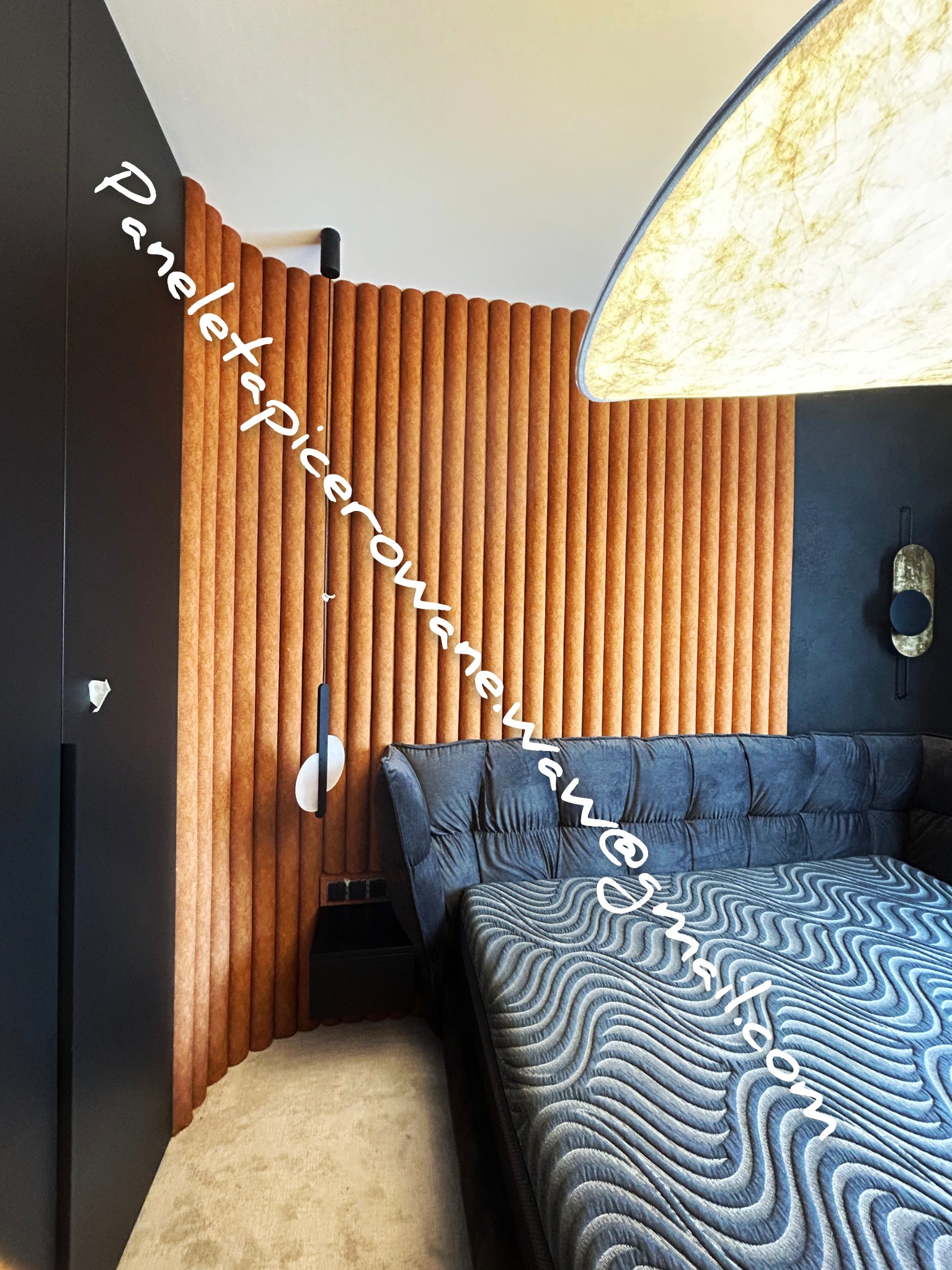 Sypialnia z łóżkiem tapicerowanym w odcieniach szarości, ceglanymi panelami ściennymi w kształcie półwałków i nowoczesną lampą wiszącą z abażurem o fakturze papieru.