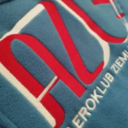 Niebieska bluza polarowa z haftowanym logo Aeroklubu Ziemi, czerwono-białe litery i samolot w prawym górnym rogu.
