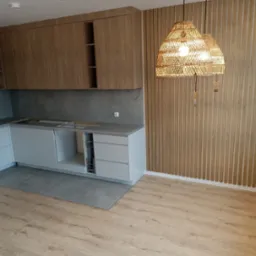 Częściowo zamontowana kuchnia z szarymi frontami i drewnianymi szafkami wiszącymi, drewniane lamele na ścianie oraz dekoracyjne lampy wiszące z abażurami z naturalnego materiału.