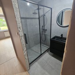 Wykończenia wnętrz - Nowoczesna łazienka w szarościach z czarną kabiną prysznicową, okrągłym lustrem i szafką pod umywalką, widok z perspektywy korytarza z surowym wykończeniem ościeżnicy.