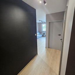 Wykończenia wnętrz - Korytarz w mieszkaniu po remoncie: czarna ściana kontrastuje z jasną podłogą z paneli, widoczne białe drzwi z czarną klamką i oświetlenie sufitowe.