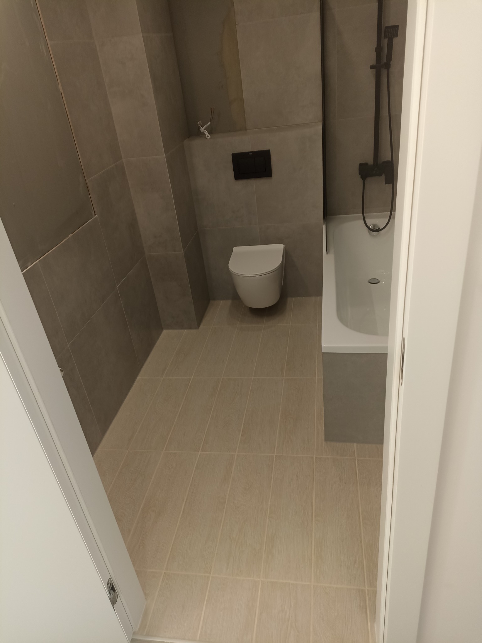 Nowoczesna łazienka w szarościach z podwieszaną toaletą, czarną armaturą prysznicową i beżowymi płytkami podłogowymi, widoczna zza uchylonych białych drzwi.