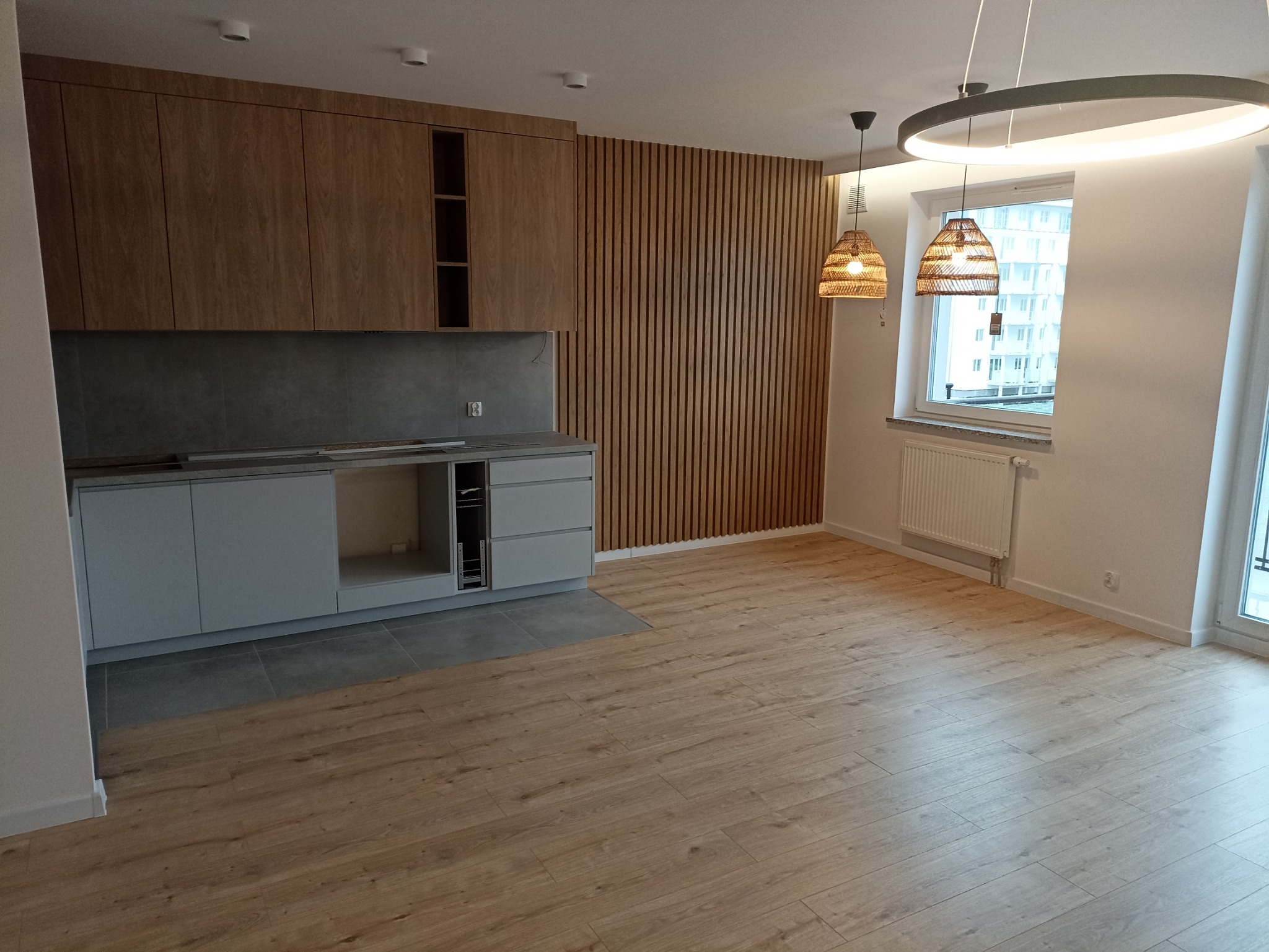 Jasne wnętrze mieszkania z drewnianą zabudową kuchenną, szarym blatem i panelami ściennymi w pionowe paski, uzupełnione oświetleniem wiszącym z abażurami z naturalnego włókna i nowoczesną lampą...