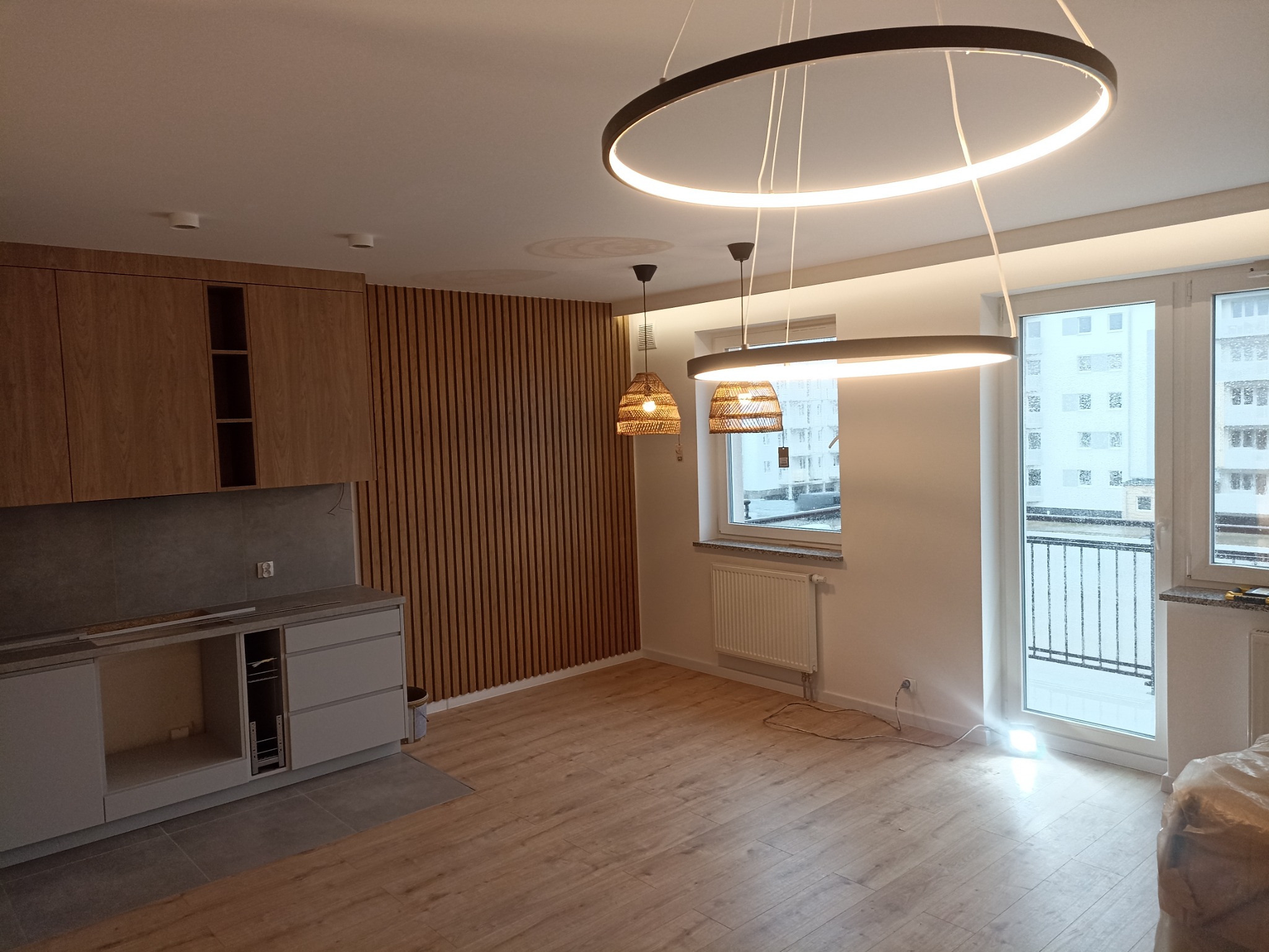 Wnętrze mieszkania po remoncie: drewniana zabudowa kuchenna, lamelowa ściana, panele podłogowe i nowoczesne okrągłe lampy sufitowe.