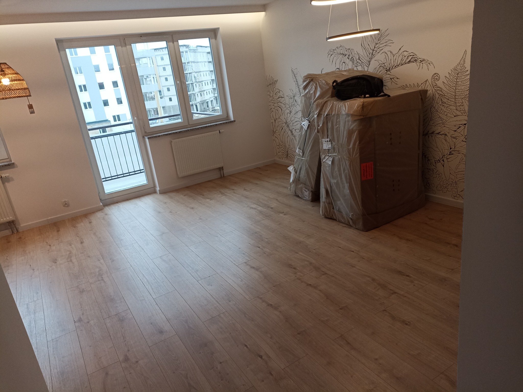 Puste mieszkanie z nową podłogą z paneli, zapakowane meble czekają na rozpakowanie, na ścianie dekoracyjny motyw roślinny.