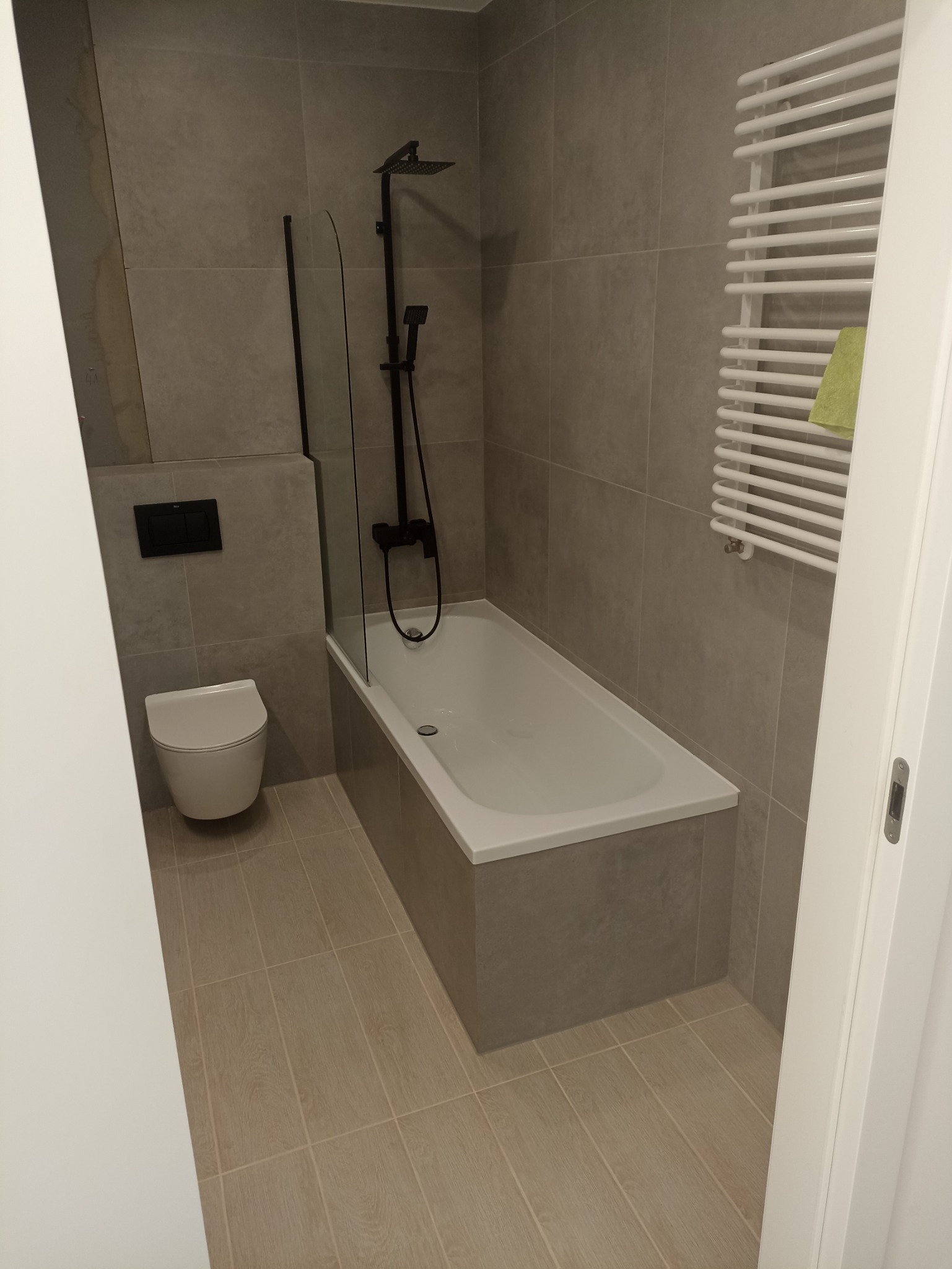 Nowoczesna łazienka w szarościach z wanną obudowaną płytkami, czarną armaturą prysznicową, wiszącą toaletą i białym grzejnikiem drabinkowym z zielonym ręcznikiem.