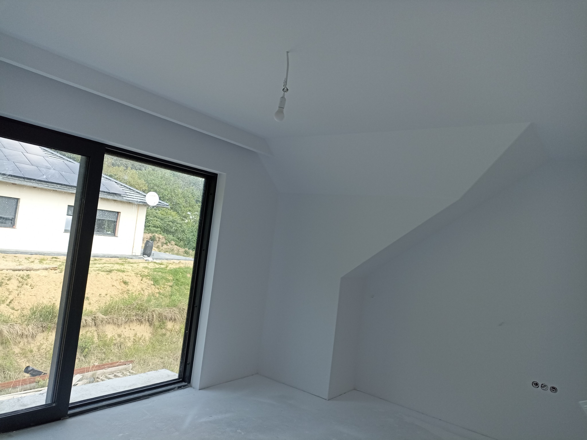 Surowe wnętrze poddasza z oknem przesuwnym, widoczne wyprowadzenia elektryczne i sufit ze skosem, w tle dom z panelami słonecznymi.