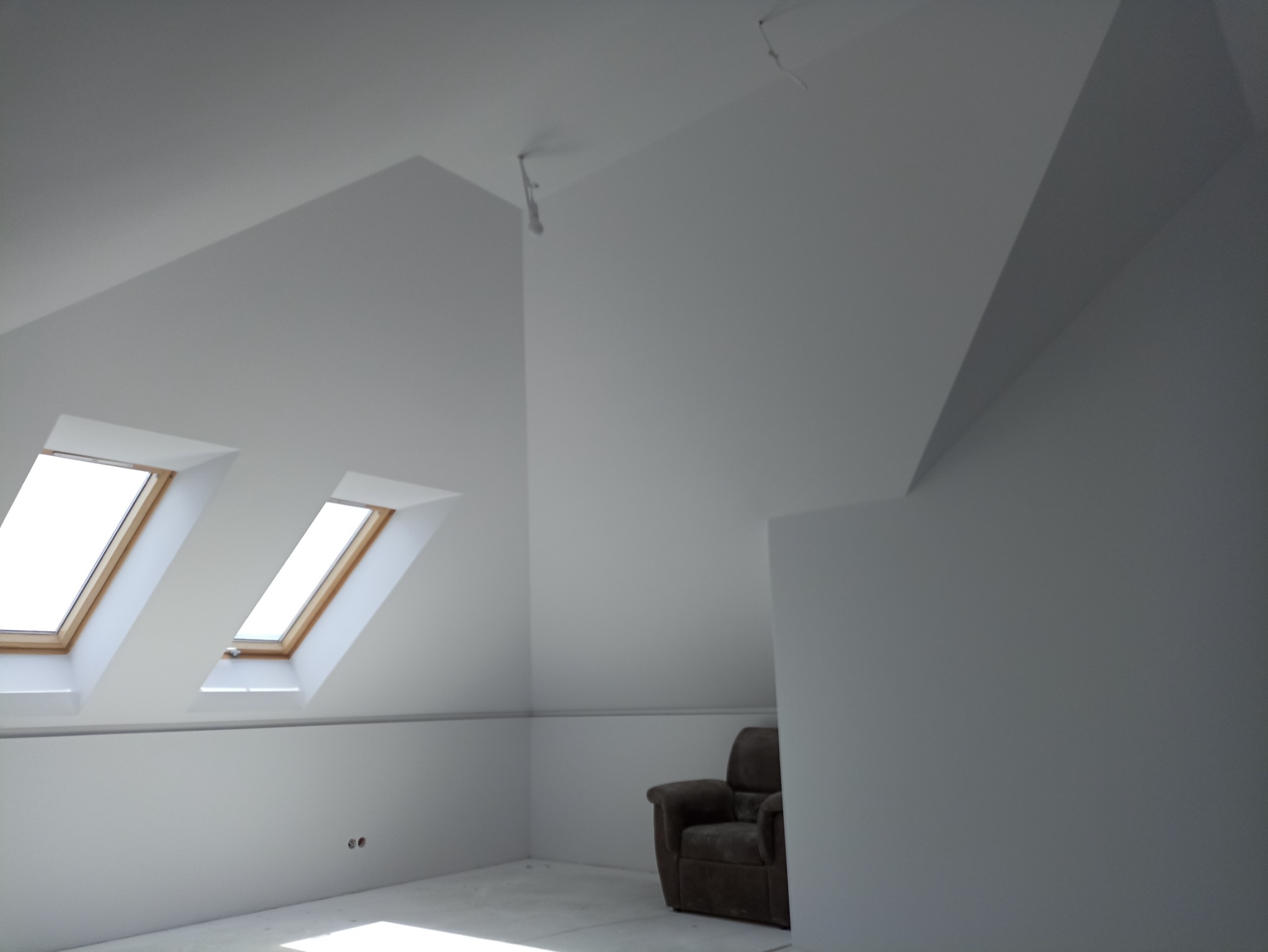 Poddasze z dwoma oknami dachowymi, świeżo pomalowane na biało, z widocznymi kablami wystającymi z sufitu i brązowym fotelem w rogu.