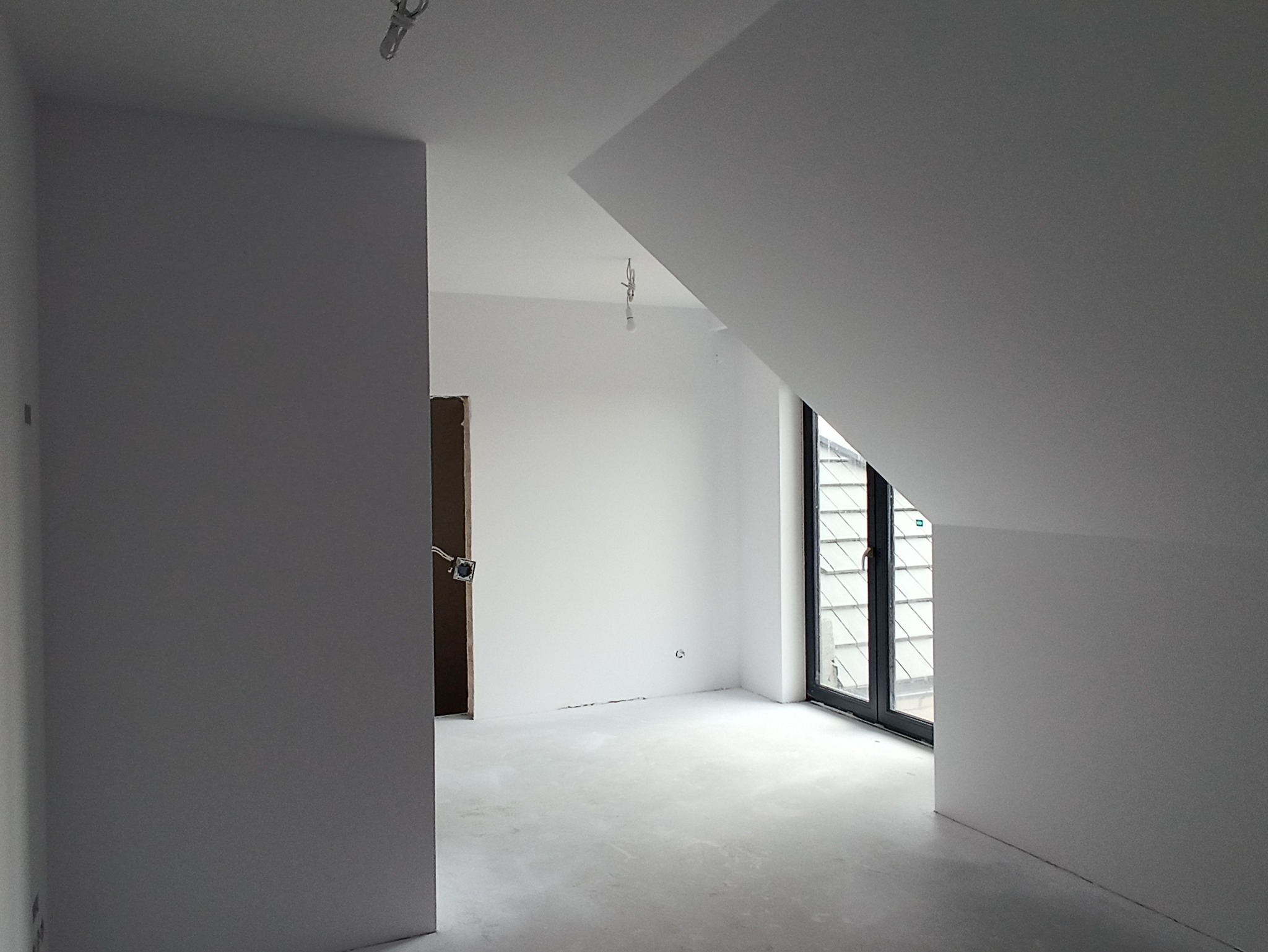 Poddasze po malowaniu na biało, widoczne okno dachowe, przygotowany otwór na drzwi i niedokończone gniazdko elektryczne na ścianie.