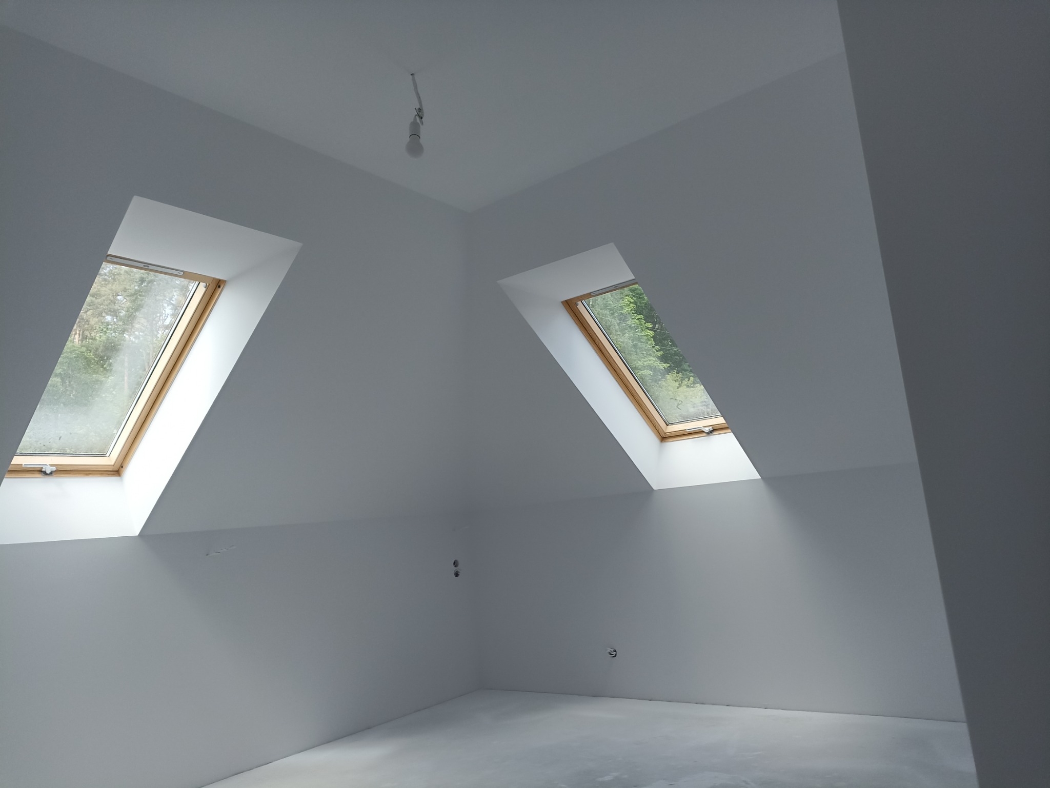 Poddasze po remoncie, świeżo pomalowane na biało, z dwoma drewnianymi oknami dachowymi, widok na zieleń za oknem, surowy beton na podłodze, widoczne punkty elektryczne na ścianie.