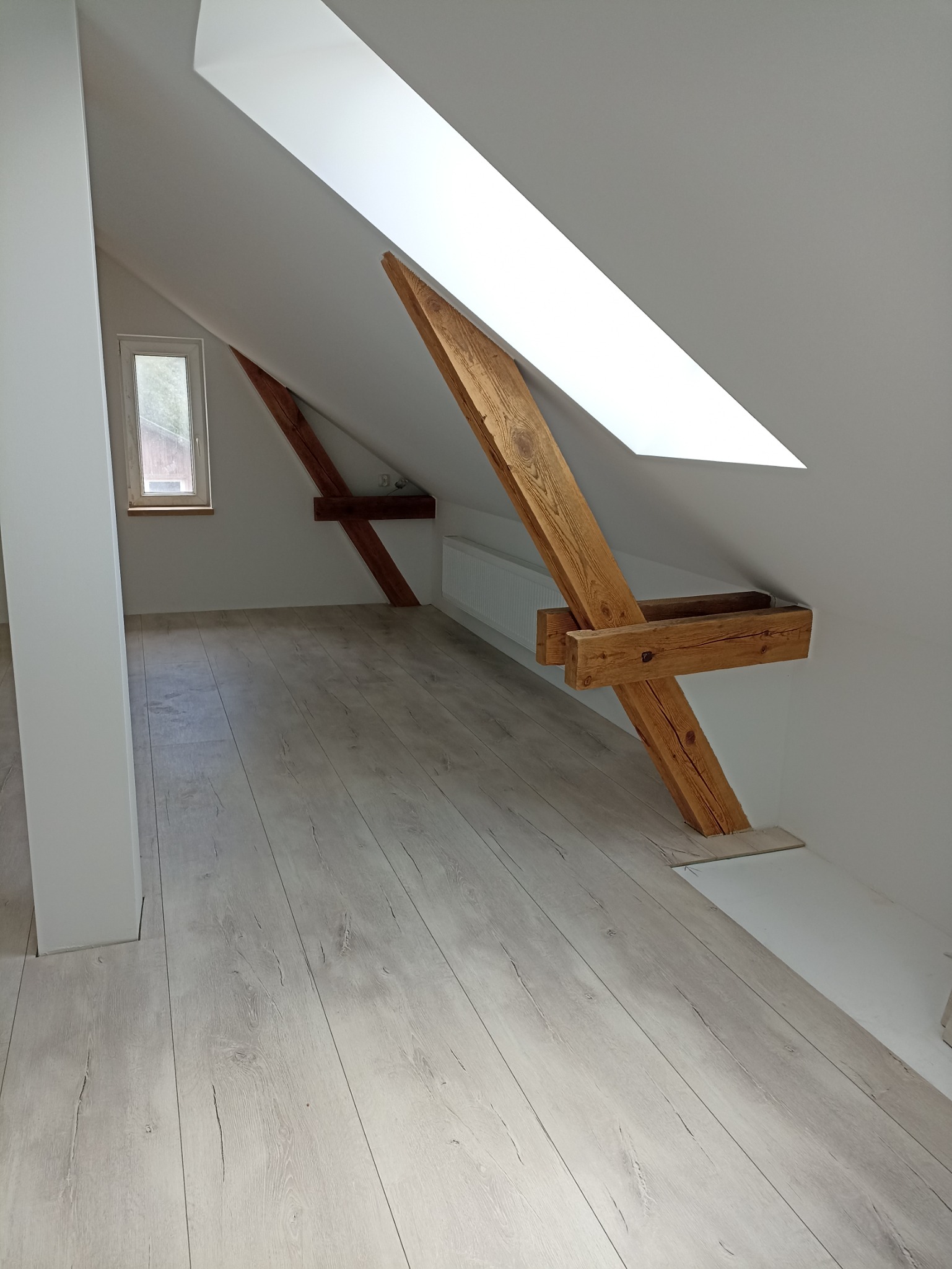 Poddasze po remoncie z jasnymi panelami podłogowymi imitującymi drewno, widoczne drewniane belki konstrukcyjne, okno i grzejnik.