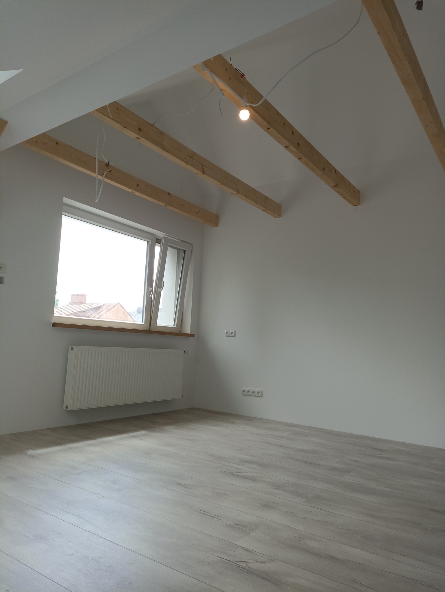 Poddasze po remoncie z jasnymi panelami podłogowymi, drewnianymi belkami stropowymi, grzejnikiem pod oknem i widoczną instalacją elektryczną.