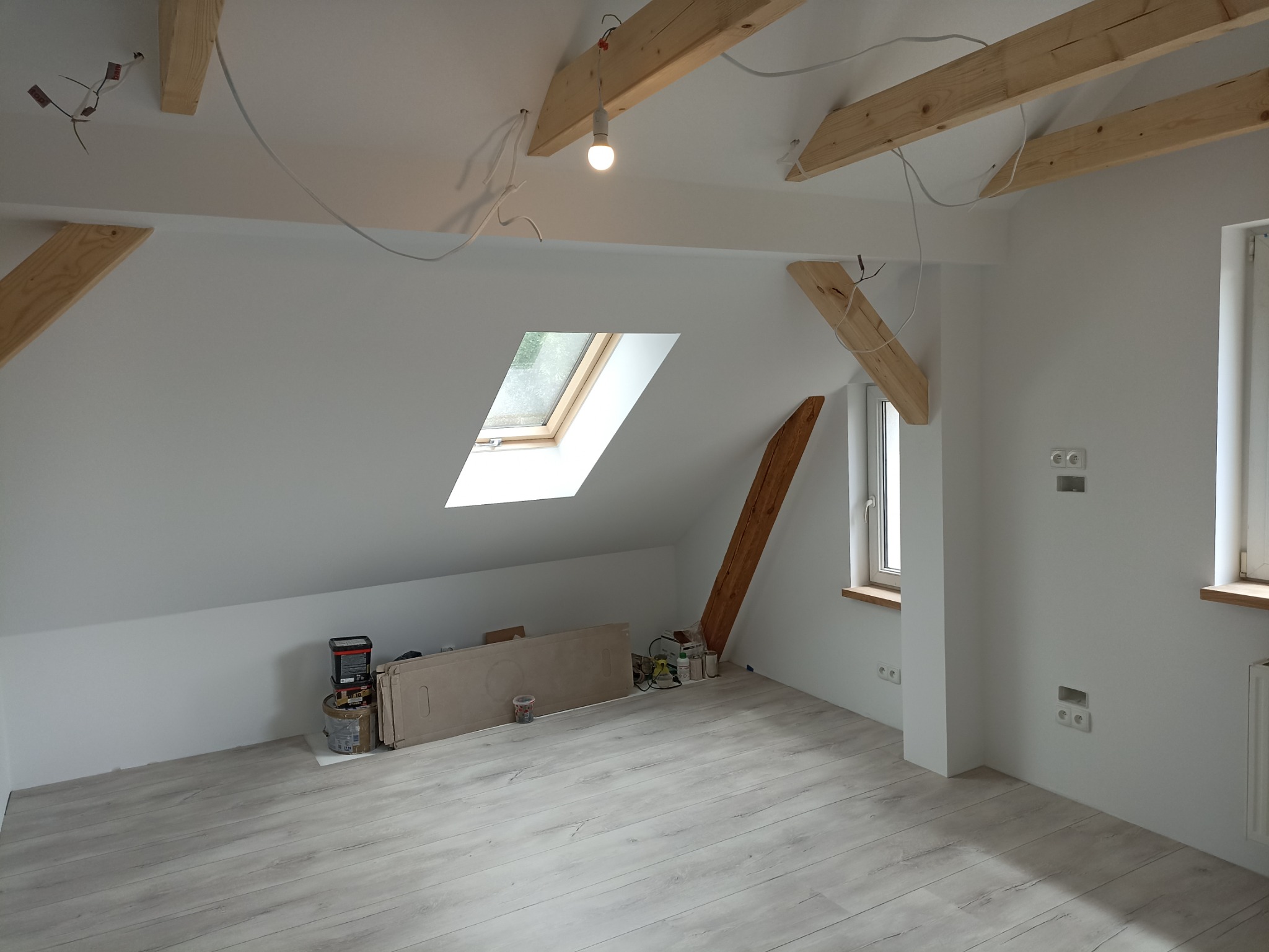 Poddasze po remoncie z drewnianymi belkami stropowymi, oknem dachowym i panelami podłogowymi w odcieniach szarości, widoczne puszki farb i narzędzia malarskie.