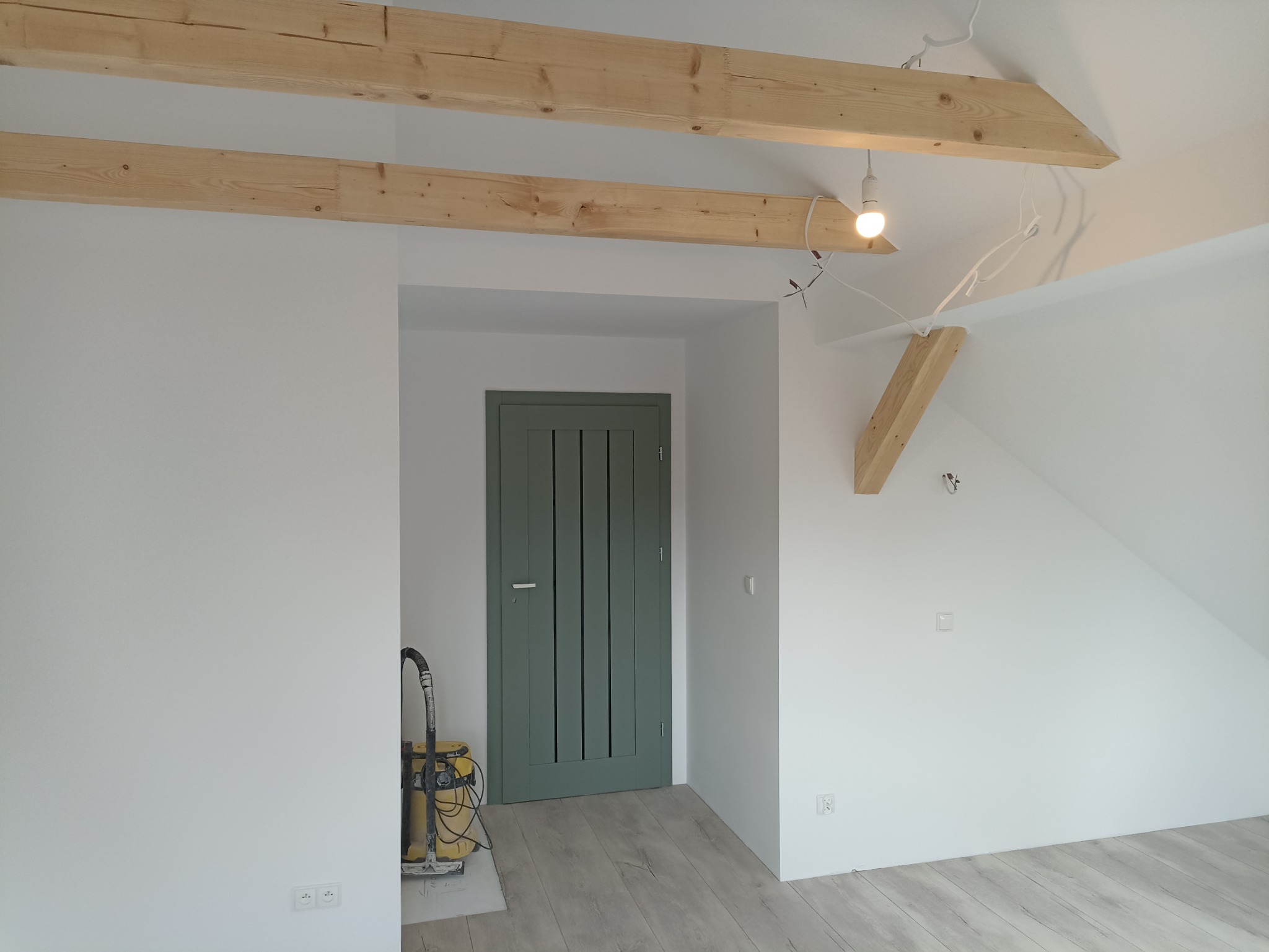 Wnętrze poddasza po remoncie: widoczne drewniane belki stropowe, nowa zielona stolarka drzwiowa, podłoga z paneli oraz odkurzacz budowlany w rogu pomieszczenia.