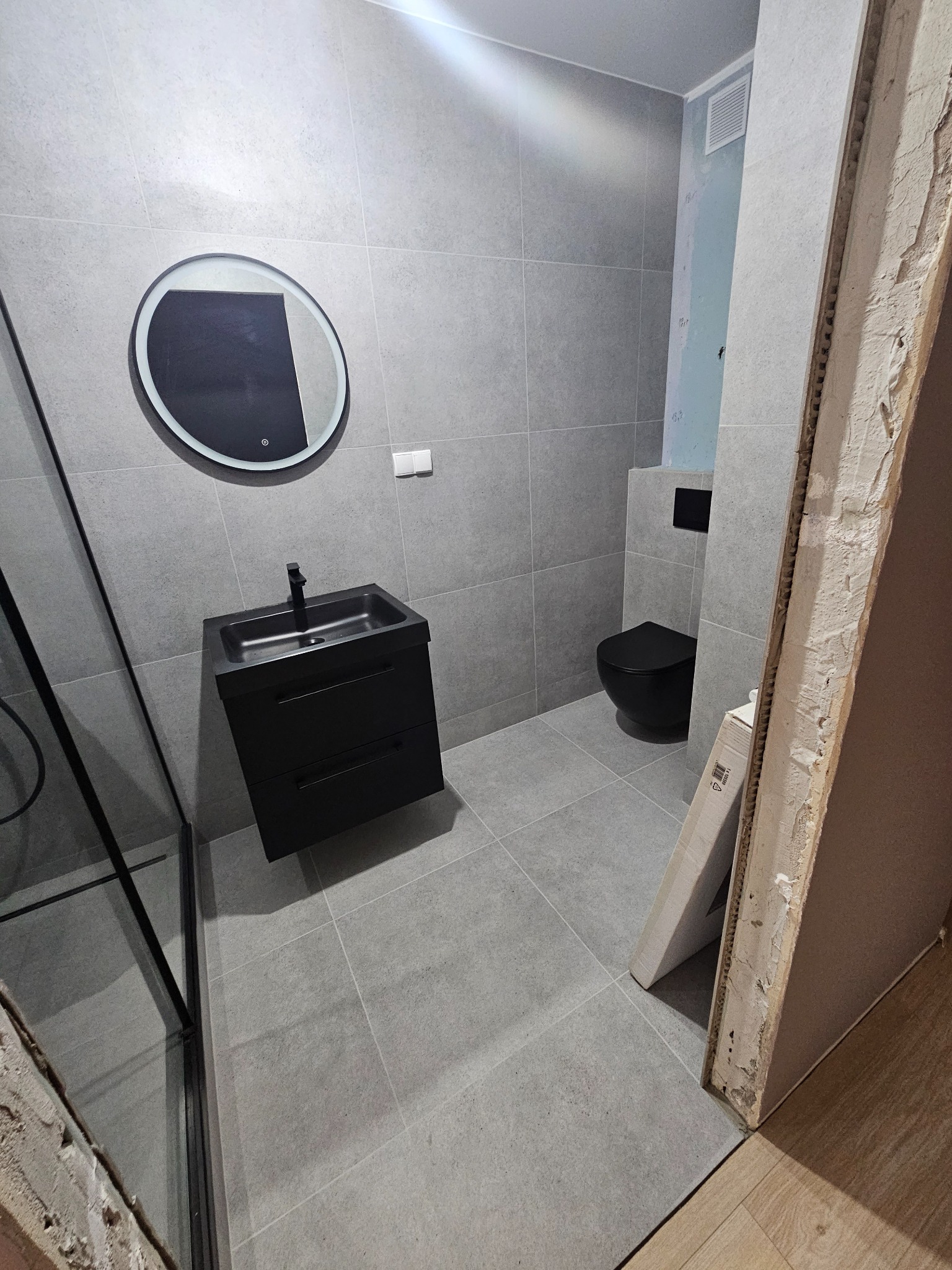 Wykończona łazienka w szarościach: czarna umywalka z szafką, okrągłe lustro z podświetleniem LED i czarna toaleta wisząca, widoczny fragment niedokończonej ściany z płyty kartonowo-gipsowej.