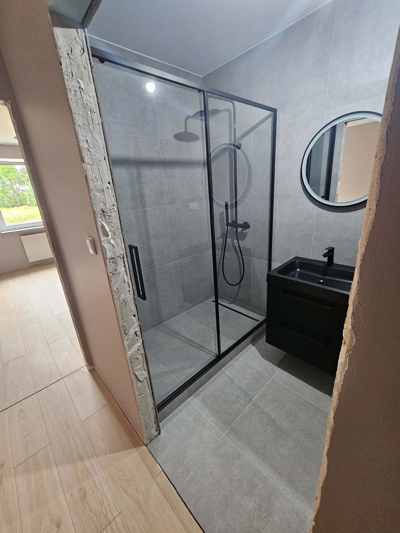 Nowoczesna łazienka w szarościach z czarną kabiną prysznicową, okrągłym lustrem i szafką pod umywalką, widok z perspektywy korytarza z surowym wykończeniem ościeżnicy.