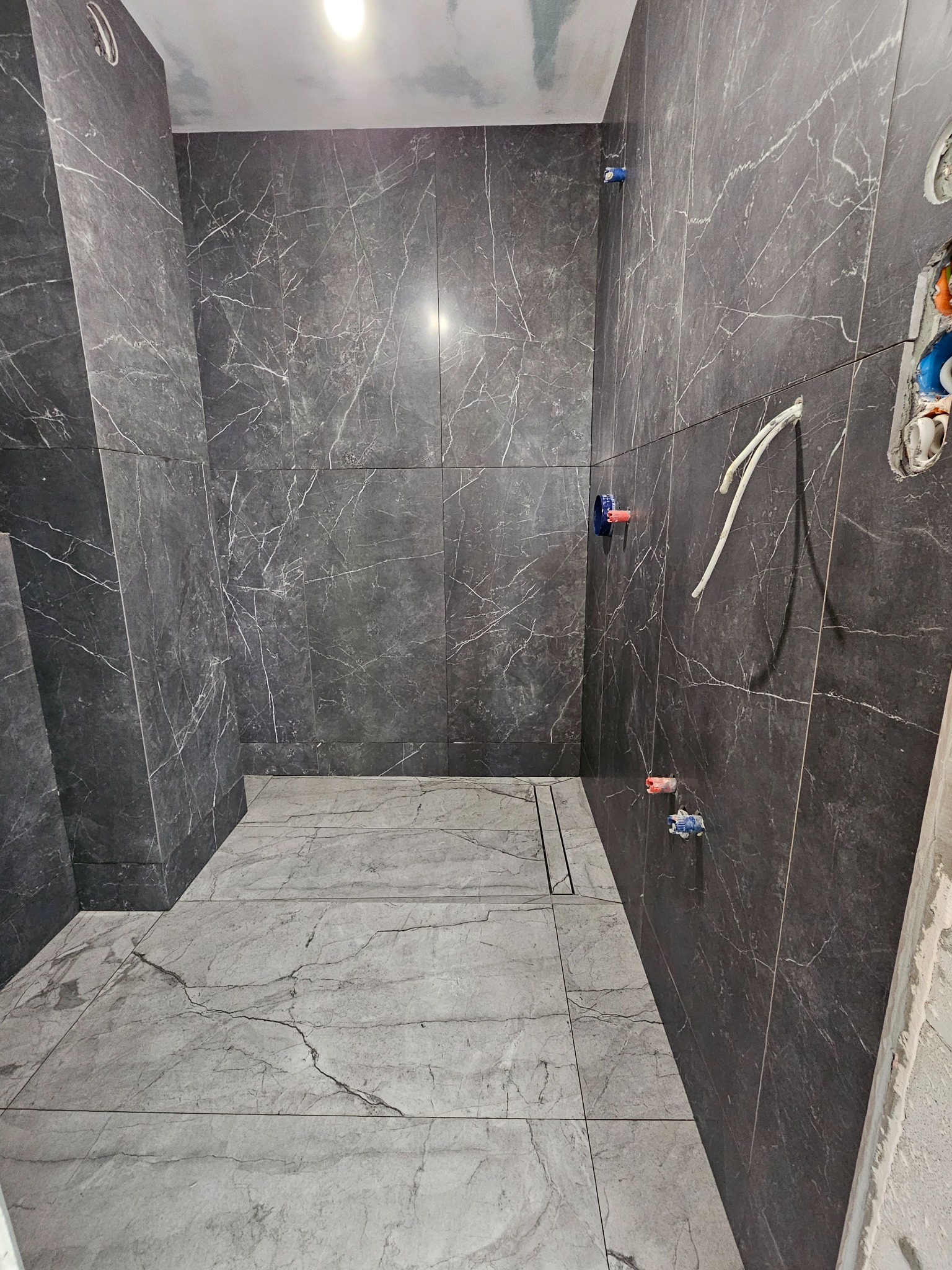 Wnętrze łazienki w trakcie wykańczania, widoczne szare płytki imitujące marmur na ścianach i podłodze, z odpływem liniowym w podłodze, oraz wystające rury instalacji wodnej.