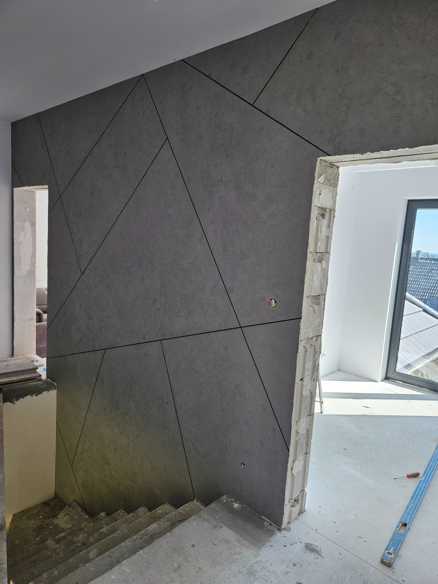 Ściana wykończona szarymi panelami o geometrycznym wzorze, widok na surowe schody i otwarte przejście do jasnego pomieszczenia z oknem.