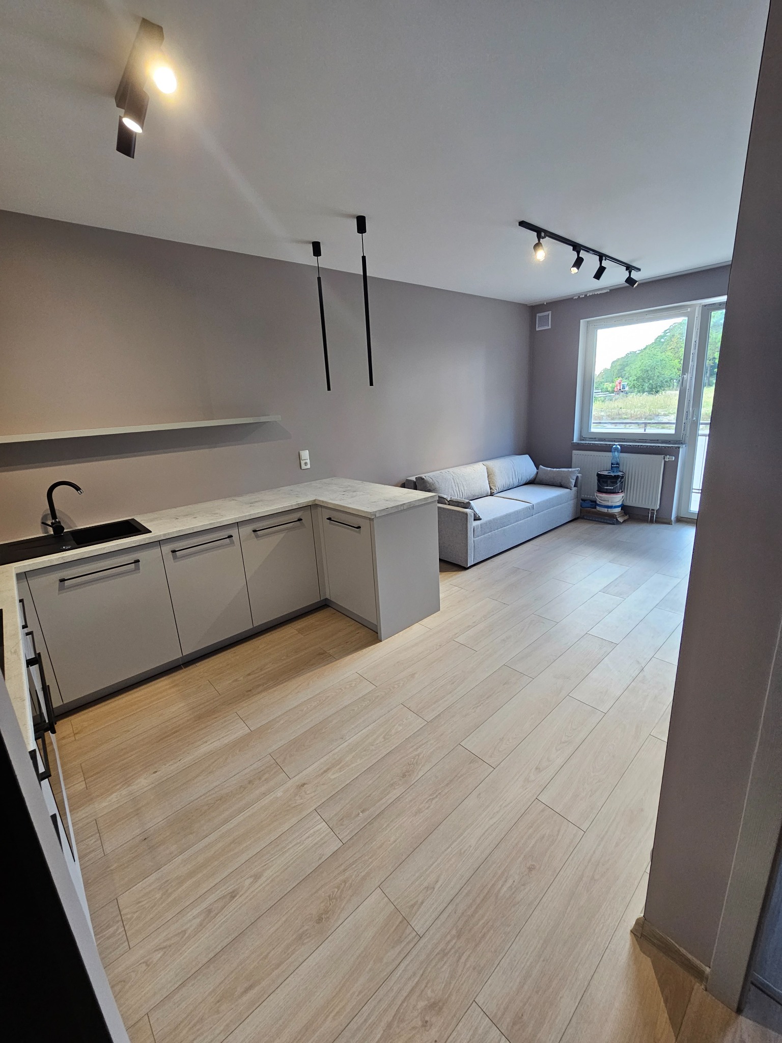 Jasne panele podłogowe w nowocześnie urządzonym pokoju z szarą kuchnią, sofą i oknem z widokiem na zieleń.