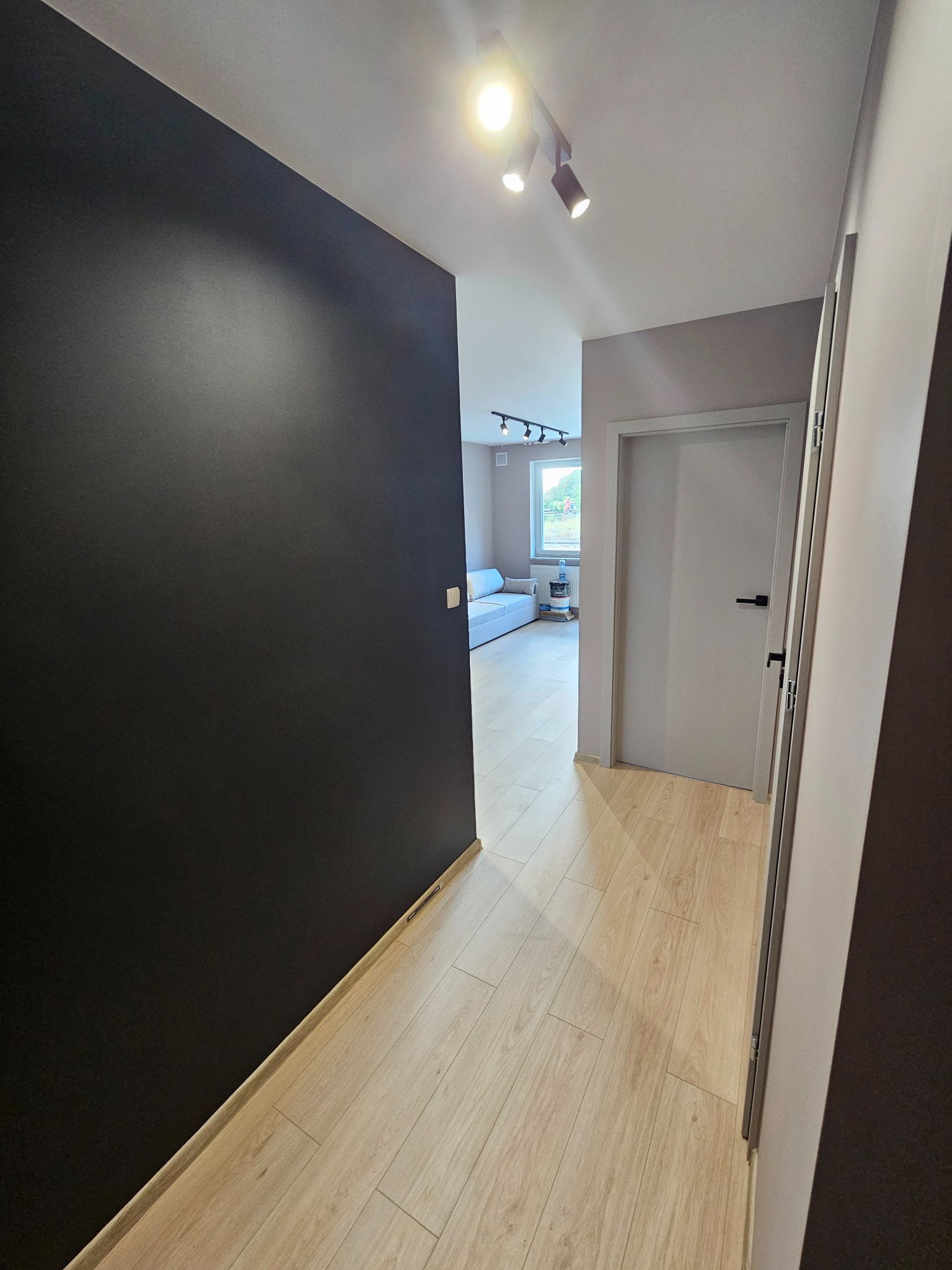 Korytarz w mieszkaniu po remoncie: czarna ściana kontrastuje z jasną podłogą z paneli, widoczne białe drzwi z czarną klamką i oświetlenie sufitowe.