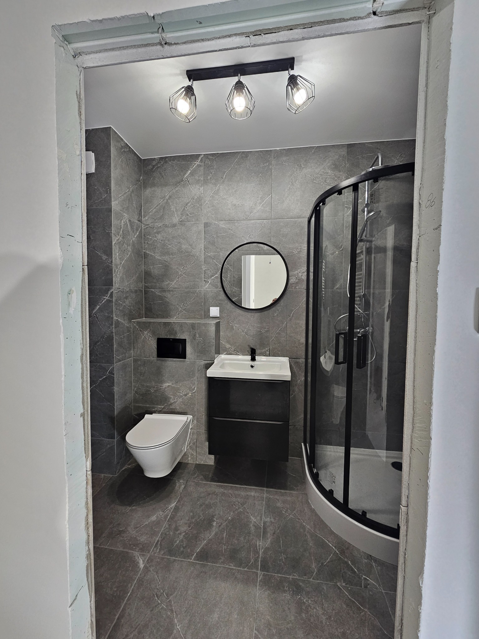 Nowoczesna łazienka z szarymi płytkami imitującymi marmur, kabiną prysznicową z czarnymi profilami, podwieszaną toaletą i okrągłym lustrem, widoczna przez surową futrynę drzwi.