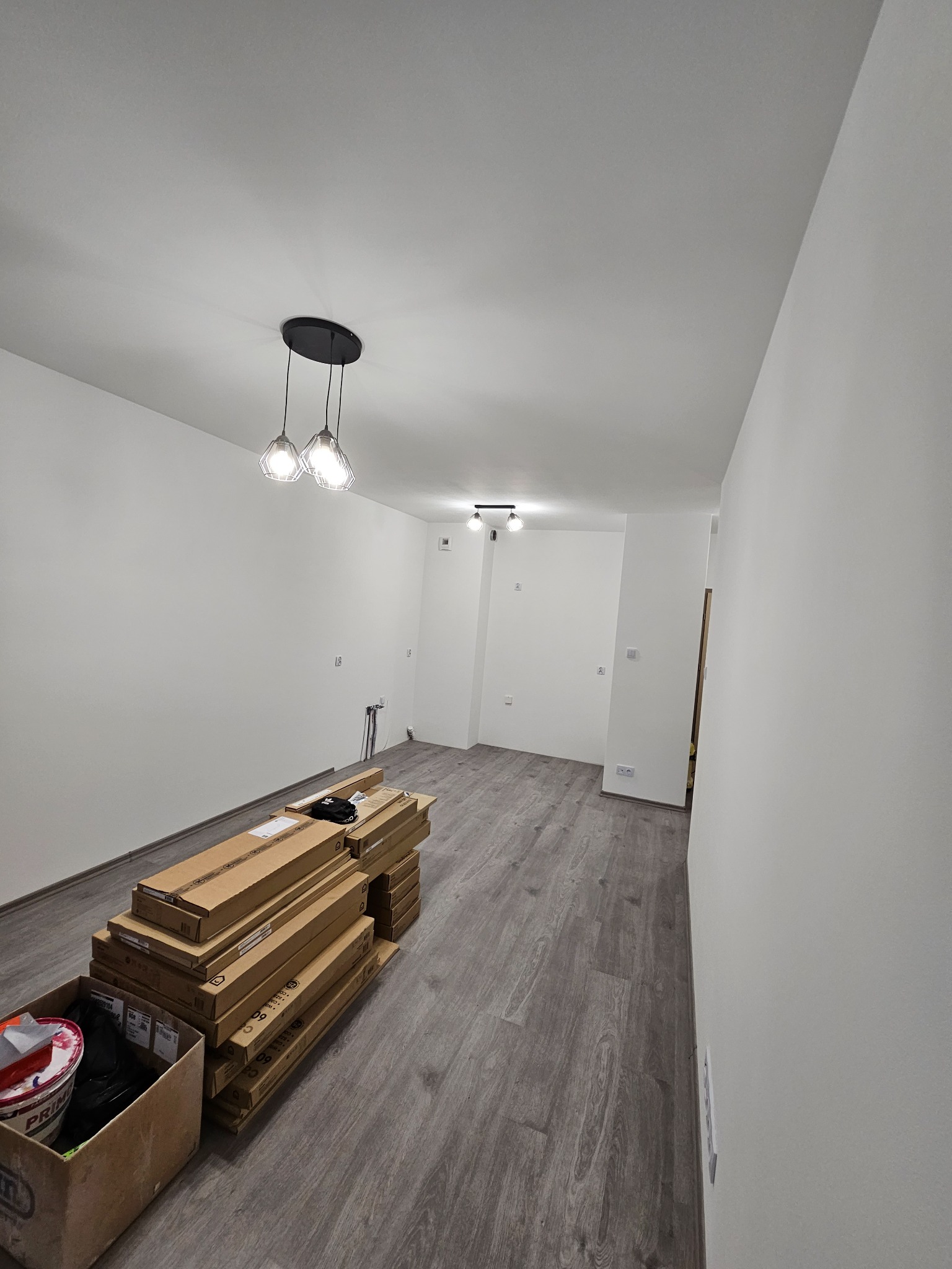 Pokój po remoncie z szarymi panelami podłogowymi, stertą kartonów z panelami w rogu, białymi ścianami i nowoczesnym oświetleniem sufitowym.