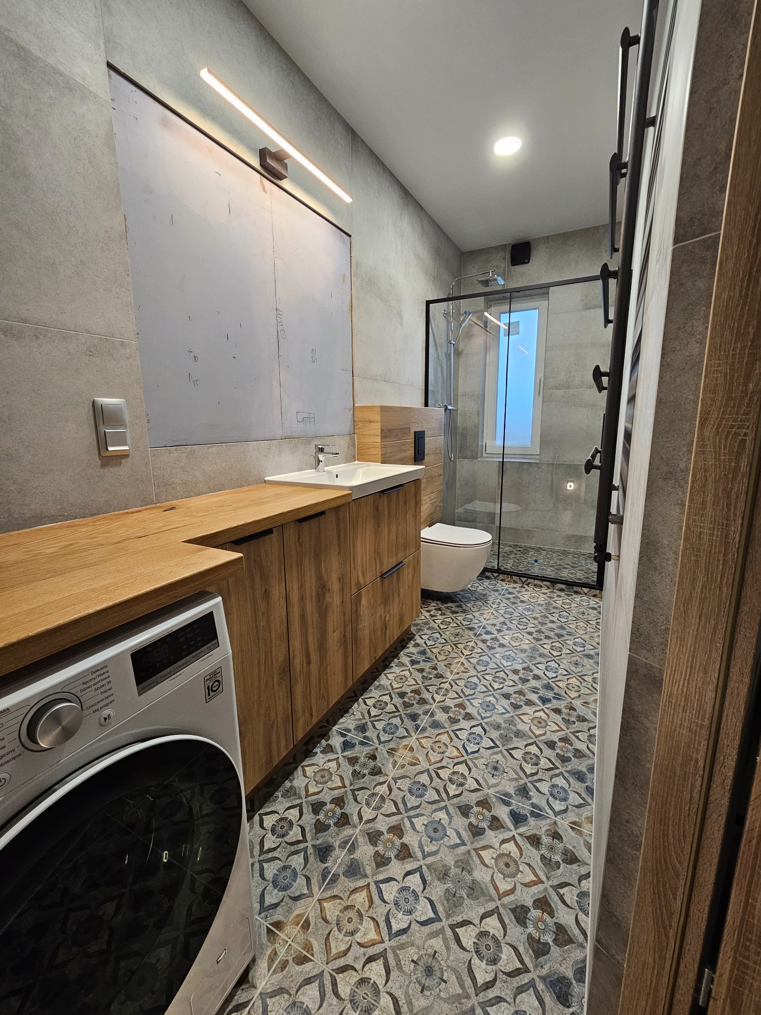 Nowoczesna łazienka z drewnianymi szafkami, umywalką, kabiną prysznicową z czarnymi ramami i pralką pod blatem. Na podłodze wzorzyste płytki w stylu vintage.
