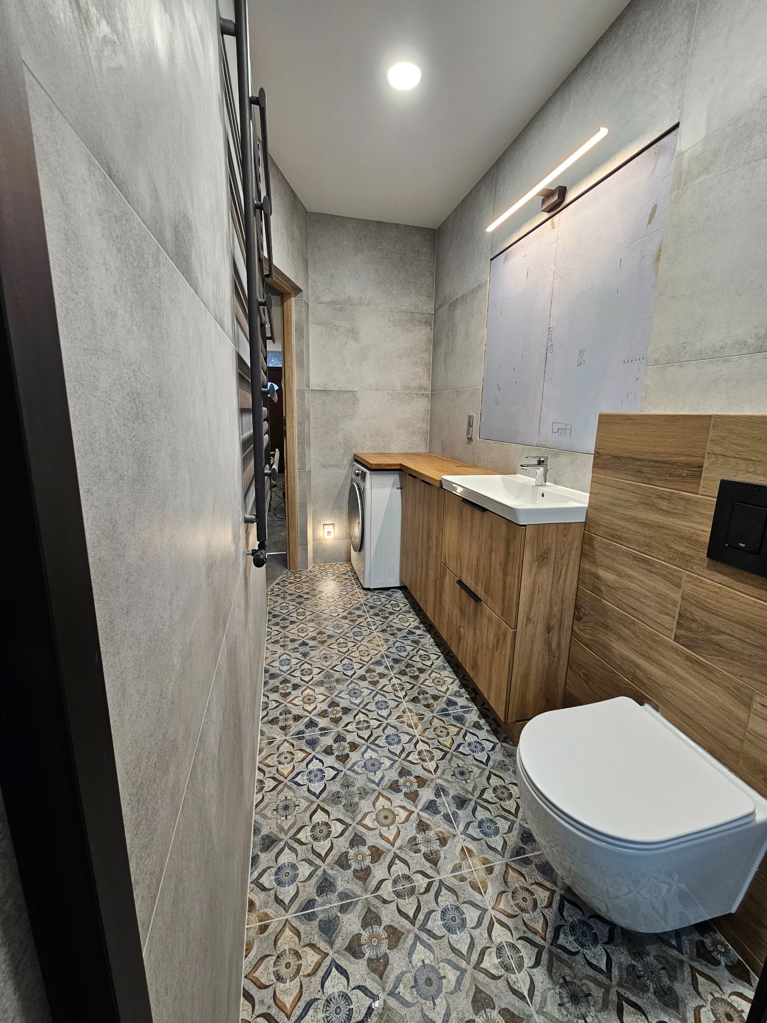 Wykończona łazienka z płytkami imitującymi drewno na ścianie, wzorzystą podłogą, umywalką w szafce z drewnianym blatem, wiszącą toaletą i grzejnikiem drabinkowym.
