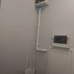 Biała skrzynka rozdzielcza elektryczna z rurą PCV na ścianie, poniżej router Wi-Fi z dwiema antenami, obok mała szafka z drzwiczkami.