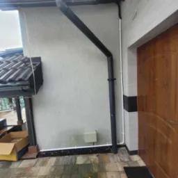 Wykończona elewacja domu z zamontowanym systemem rynnowym w kolorze grafitowym, widoczna skrzynka elektryczna i fragment instalacji pod rynnami.