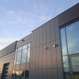 Zewnętrzny widok budynku z szarej blachy z zamontowanymi kamerami monitoringu i czujnikiem alarmowym, okna odbijają błękitne niebo.