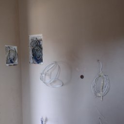 Tynki maszynowe - Świeżo otynkowana ściana w rogu pomieszczenia z widocznymi przewodami elektrycznymi wychodzącymi ze ściany i ze skrzynek elektrycznych.