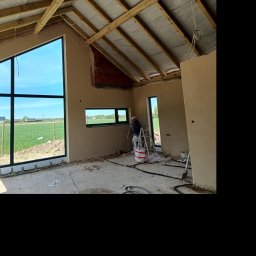 Tynki maszynowe - Prace wykończeniowe w surowym wnętrzu z dużym oknem. Widoczny pracownik na drabinie, przygotowane ściany, surowy strop i podłoga. Za oknem zielony krajobraz.