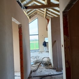 Tynki maszynowe - Wnętrze w trakcie prac: ściany pokryte świeżym tynkiem, widoczny pracownik przy oknie. Surowe wykończenie, widoczne przewody i drewniana konstrukcja dachu.