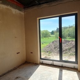 Tynki maszynowe - Wnętrze w stanie deweloperskim: ściany po tynkowaniu, widoczne puszki elektryczne, duże okno z widokiem na zewnątrz. Podłoga surowa, wylewka. Ściany w jasnym, beżowym odcieniu.