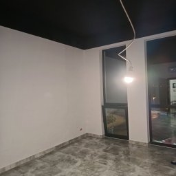 Tynki maszynowe - Wykończone wnętrze z czarnym sufitem, białymi ścianami i szarą podłogą z płytek. Widoczne okna z ciemnymi ramami i żarówka na kablu.