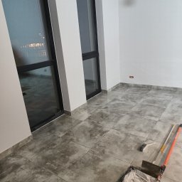 Tynki maszynowe - Pokój po malowaniu ścian na biało, widoczne narzędzia malarskie: wałek w kuwecie, pędzel i teleskopowy kij. Szare płytki na podłodze, duże okna.