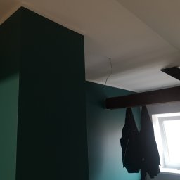 Tynki maszynowe - Ściana pomalowana na ciemny zielony kolor w pokoju na poddaszu z oknem i widoczną belką stropową. Na belce wiszą kurtki.