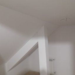 Tynki maszynowe - Białe wnętrze poddasza po remoncie, widoczne gładkie ściany, fragment grzejnika i wystające kable z sufitu. Kadrowanie od dołu, z lekkim rozmytym efektem.